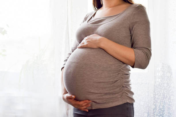 Pregnant woman with big belly at window. Maternity prenatal care and woman pregnancy concept. Young pregnant woman holds her hands on her swollen belly. Love concept. Horizontal with copy space.